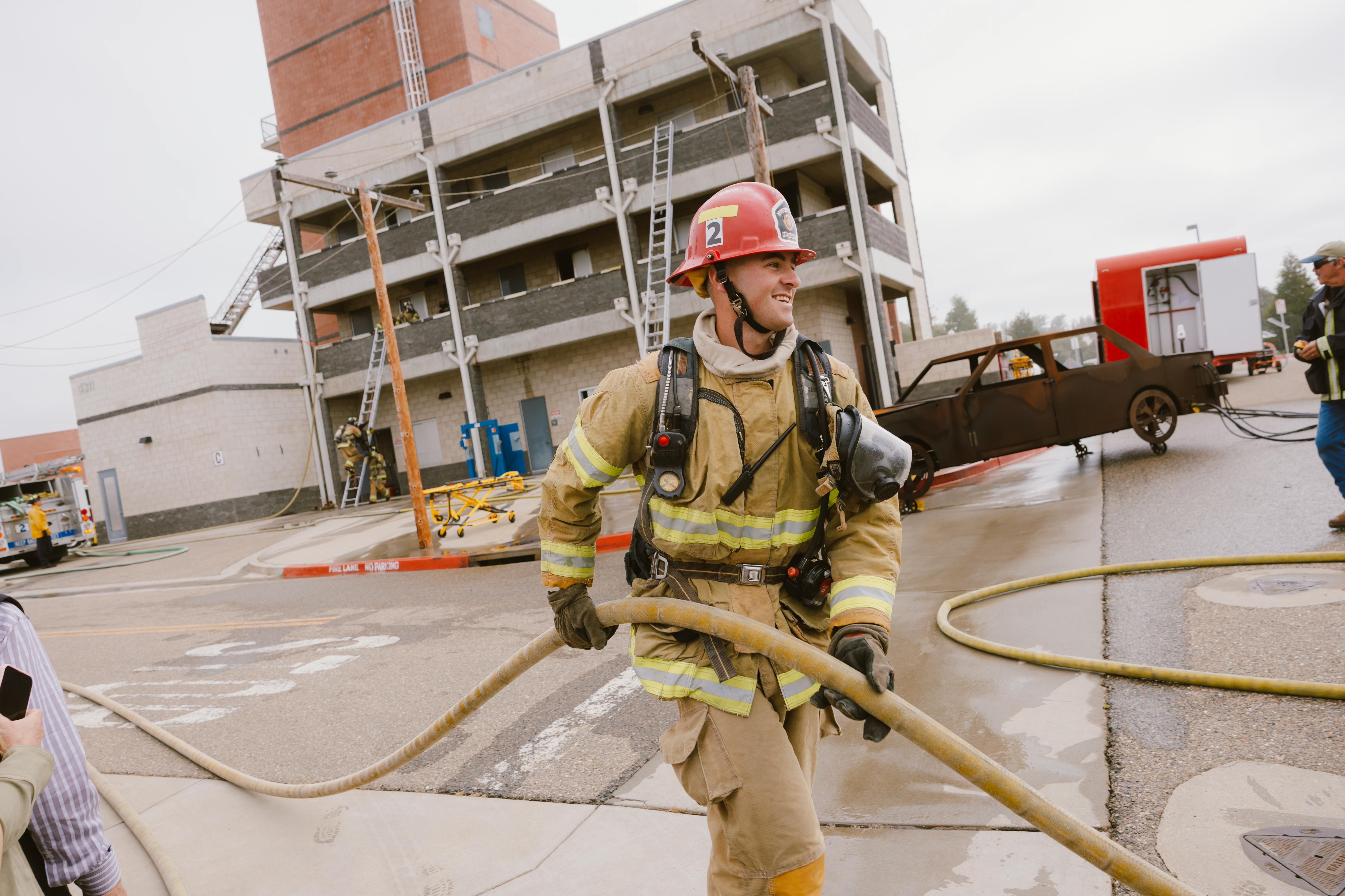 A firefighter in full protective gear carries a fire hose during a training exercise at Allan Hancock College’s Fire Academy graduation on May 15, 2026. Behind the firefighter is a multi-story fire training tower, fire equipment, and academy participants practicing emergency response skills.